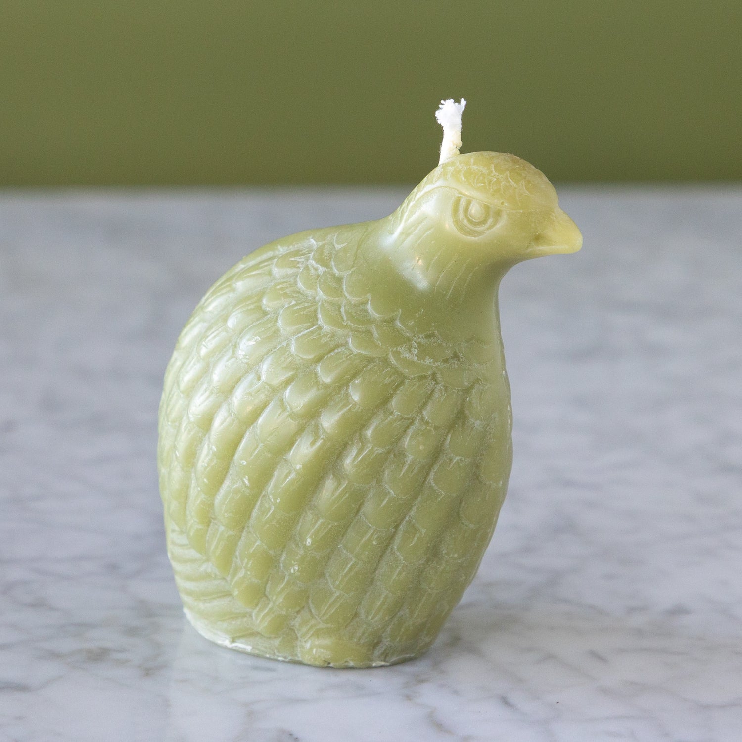 A Partridge Beeswax candle in sage on a marble surface with a green wall background.