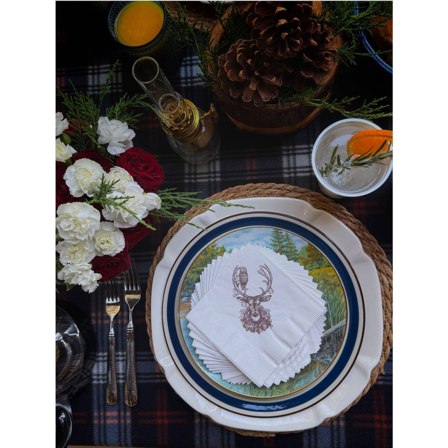 Decorative table setting with a plate, napkins with the deer design, and flowers on a plaid tablecloth.