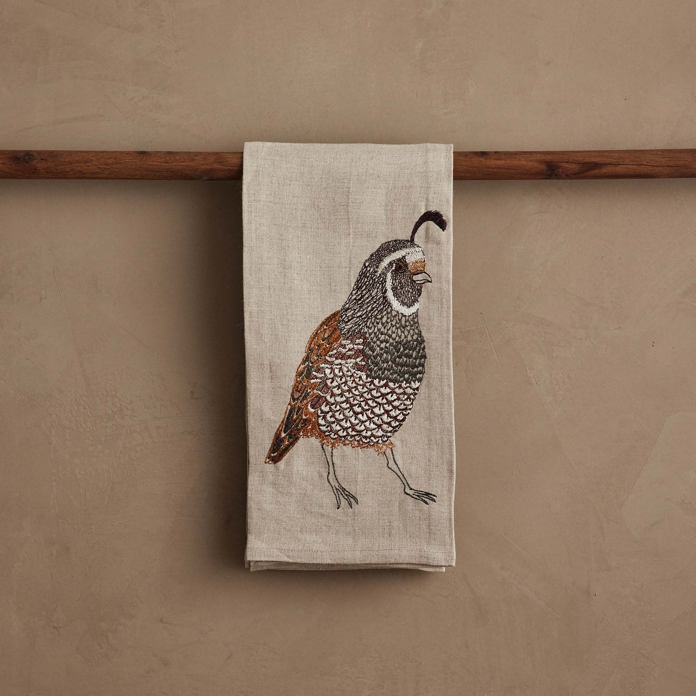 Towel with a quail design hanging on a wooden rod against a beige wall.
