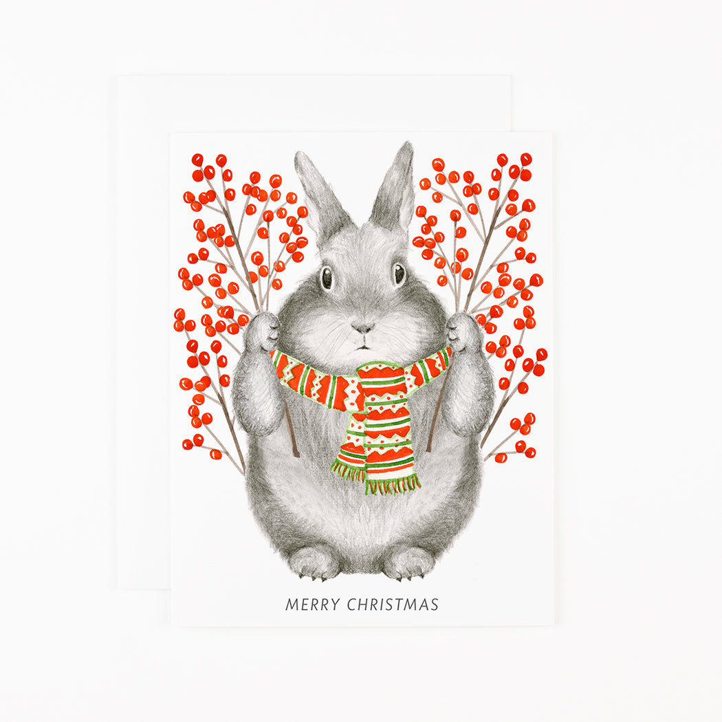 Winter Berry Bunny Card by Dear Hancock of a hand-drawn illustration of a bunny holding holding sprigs of bright red winterberry with a scarf around his neck and text below that says &quot;Merry Christmas&quot;&gt;