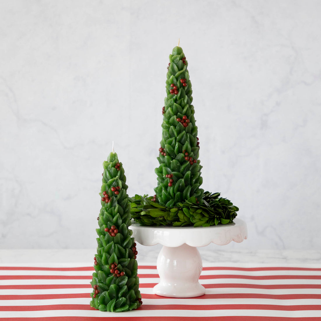 A set of two festive candles designed to resemble green Christmas trees with red berries, displayed on a white surface with a red and white striped background.