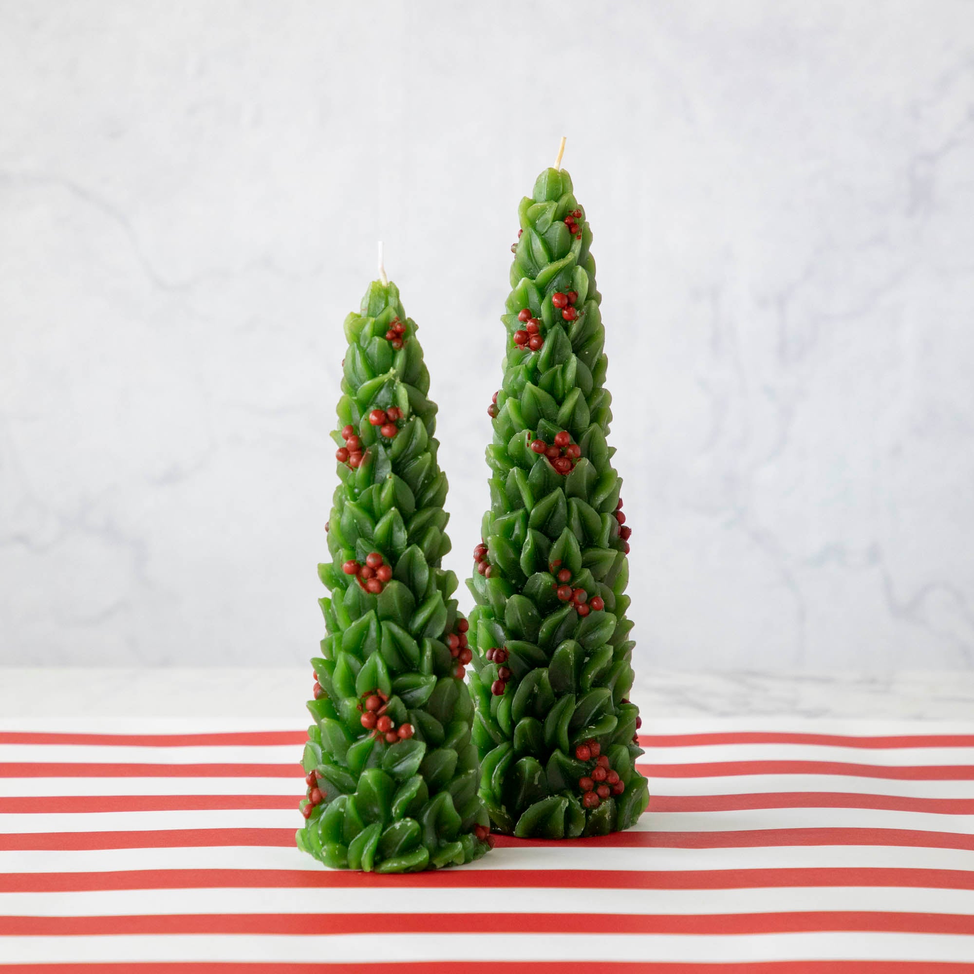 A set of two festive candles designed to resemble green Christmas trees with red berries, displayed on a white surface with a red and white striped background.
