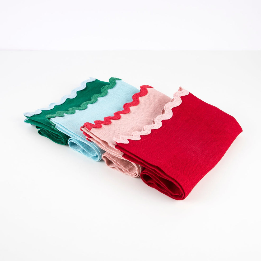 Four folded fabric napkins in green, blue, pink, and red with scalloped edges on a white background.