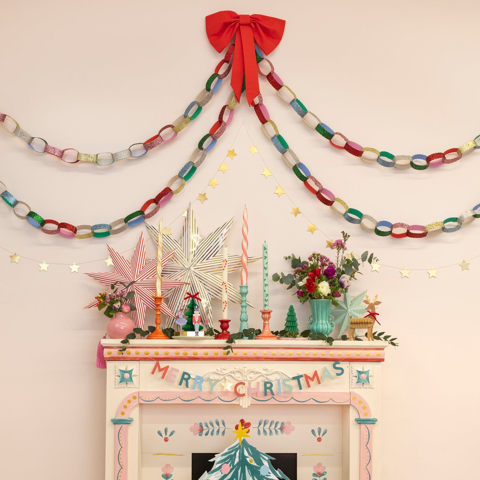 Decorative Christmas mantel with colorful garlands, candles, and festive decorations on a light pink wall.