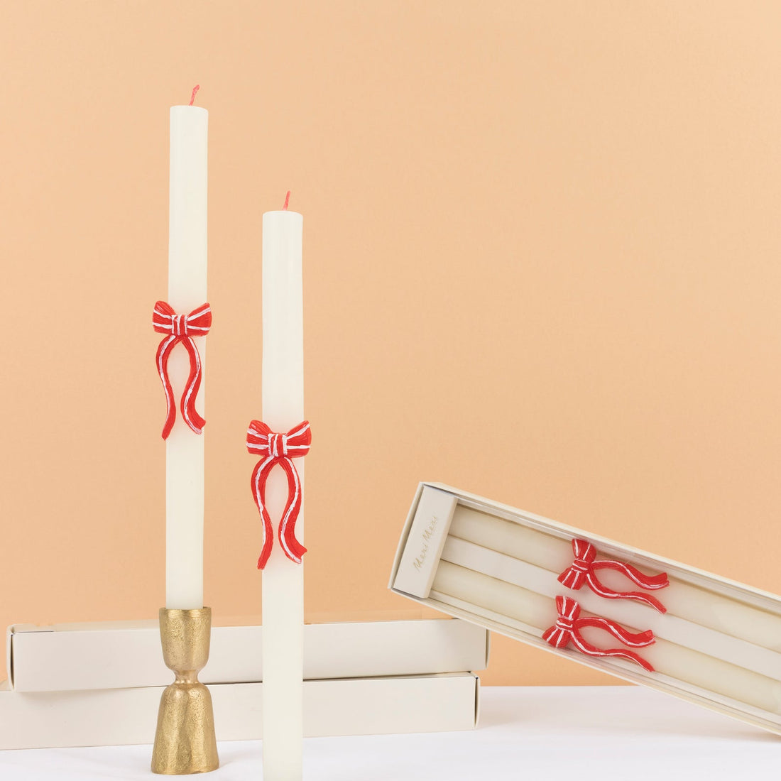 Two white candles with red bow decorations on a peach background, accompanied by a gold candle holder.