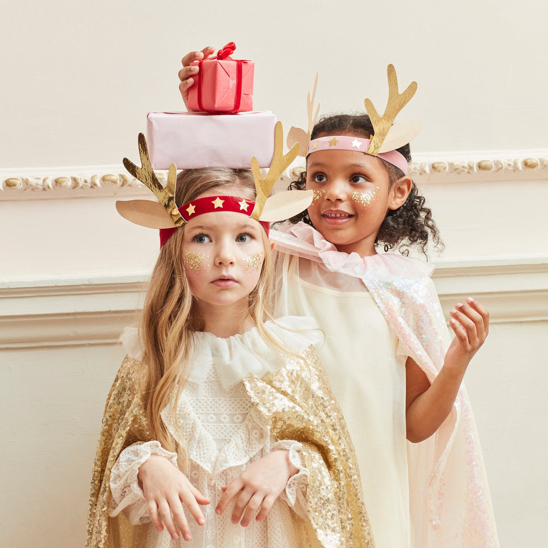 Two children wearing festive outfits with reindeer antlers and gift boxes on a decorative background.