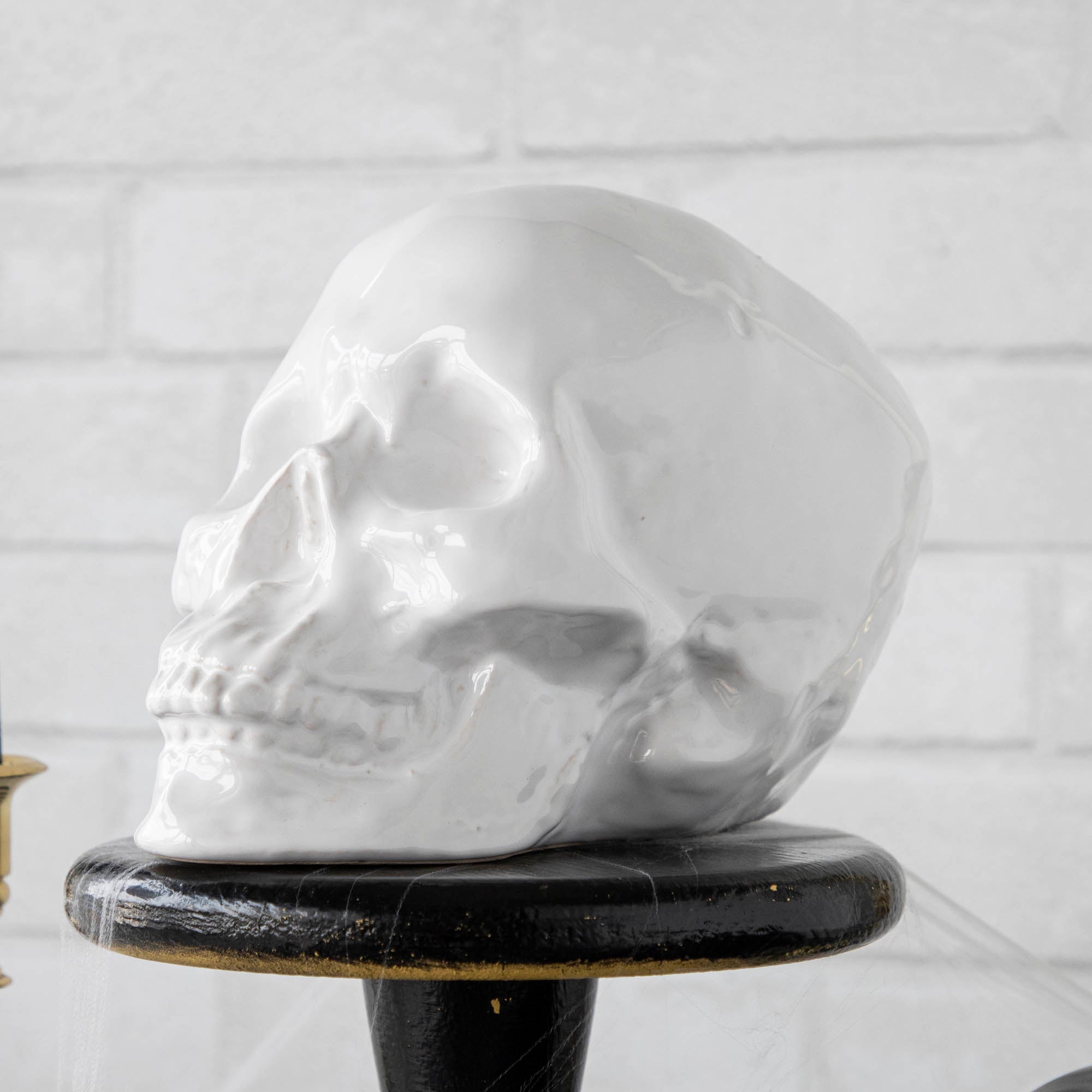 Two HomArt ceramic Skull Heads on a striped tablecloth flanked by lit candles in brass holders against a white brick background, serving as an ominous decor piece.