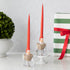 Two taper candles with Santa Claus designs on them, placed in clear candle holders, with one holder sitting on a white stand. The candles are red with white accents and Santa Claus face details.
