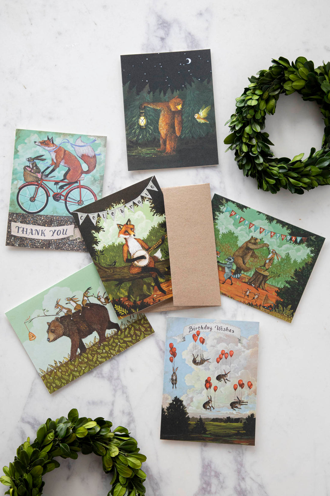A set of six different whimsically illustrated Elizabeth Foster greeting cards, featuring foxes, bears and rabbits, on a white marble table.