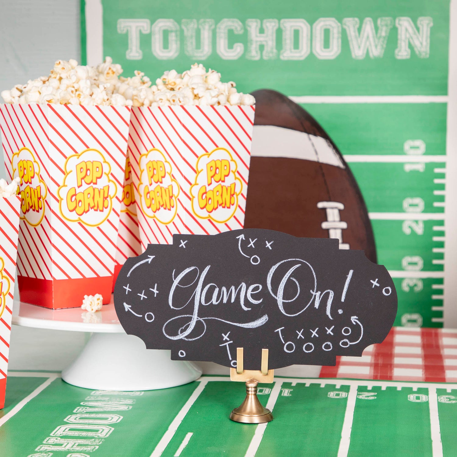 A football-themed snack table featuring a Black Frame Table Accent standing in a place card holder, with "Game On!" hand-written in white script along with accents of Xs and Os with arrows in the style of football strategy maps.