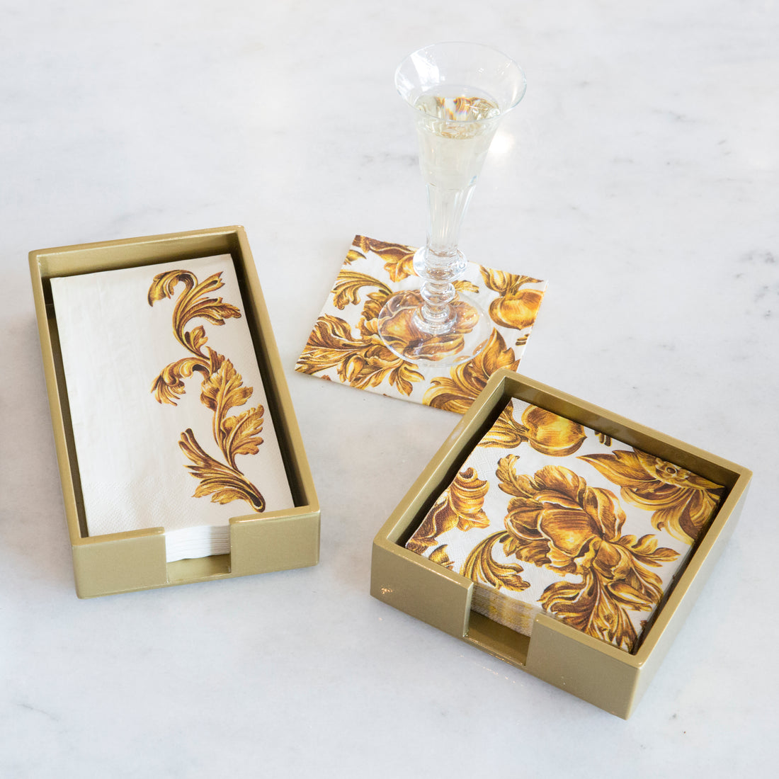 A gold lacquered-wood holder for beverage napkins, displayed alongside a glass of champagne and napkins with a leafy, gold design.