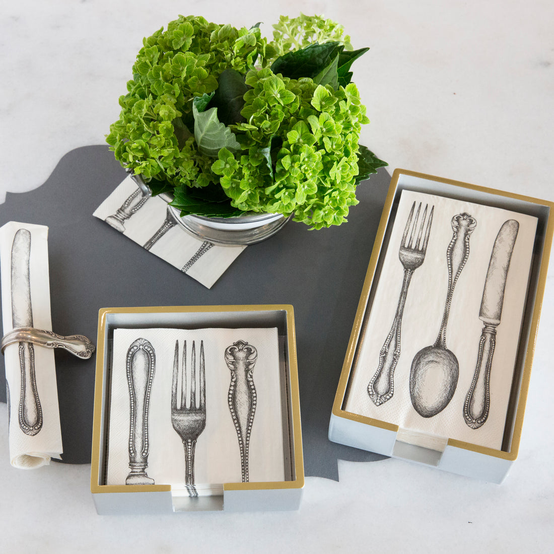 A lacquered wood napkin holder with silver and gold trim, decorated with illustrations of cutlery, displayed on a table with a vase of flowers in the background.