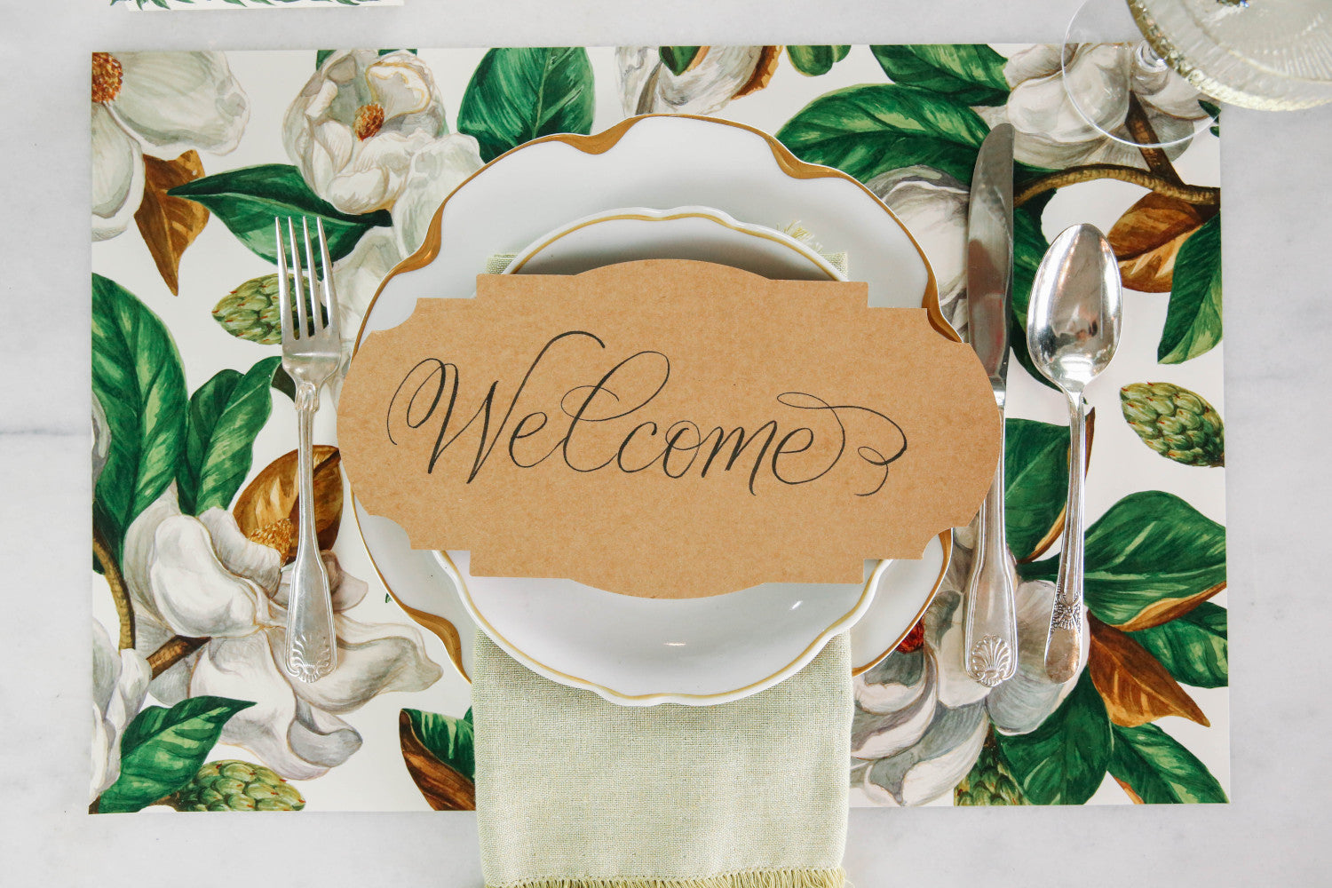 An elegant floral place setting featuring a Kraft Frame Table Accent resting on the plate with "Welcome" written in beautiful script.