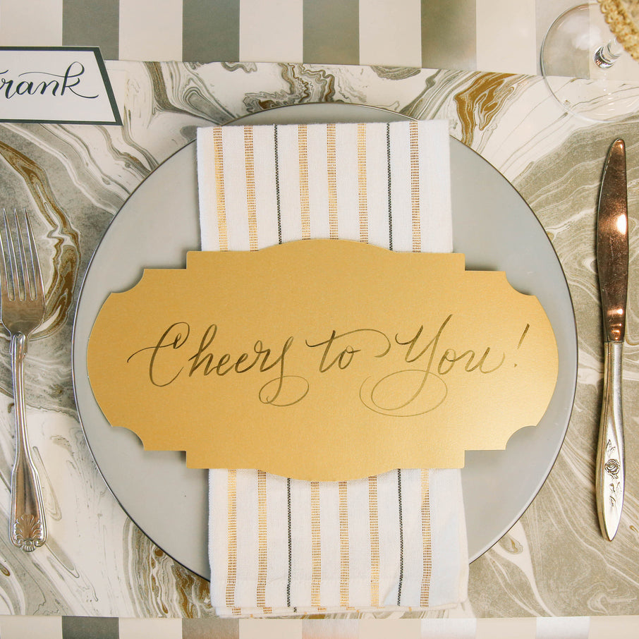 Top-down view of an elegant place setting featuring a Gold Frame Table Accent resting on the plate with &quot;Cheers to You!&quot; written in beautiful script.