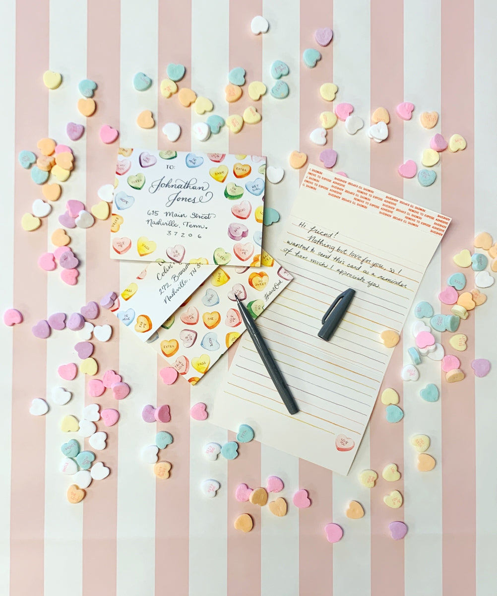 A Conversation Hearts Fold Note with confetti and a pen on a striped background, by Hester &amp; Cook.