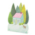 A die-cut freestanding place card featuring a whimsical illustration of a teal and pink house in a snowy field surrounded by tall green trees and a single deer, with the blank snow-covered front yard serving as a blank area for personalization.