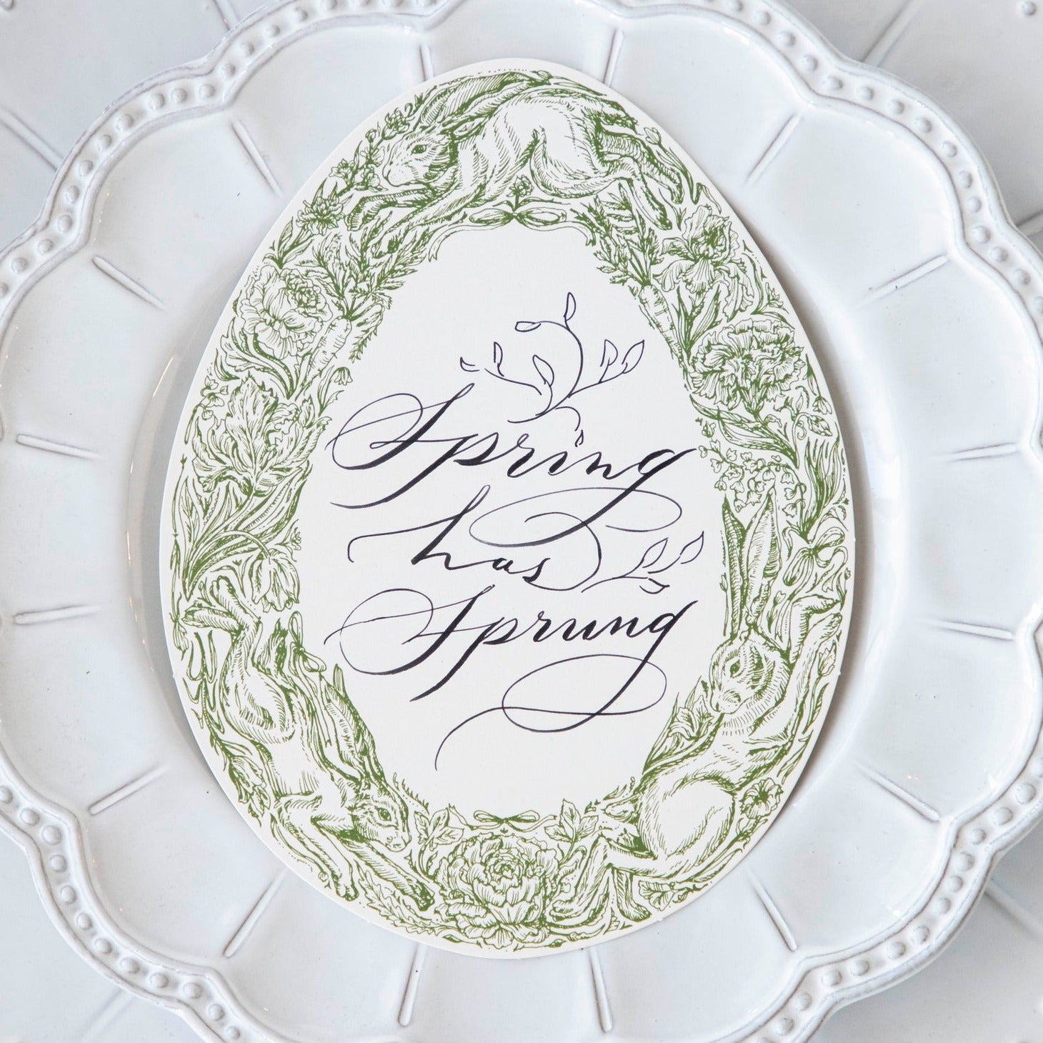 Close-up of a Greenhouse Hares Table Card with "Spring has Sprung" written on it in elegant script resting on a white plate.