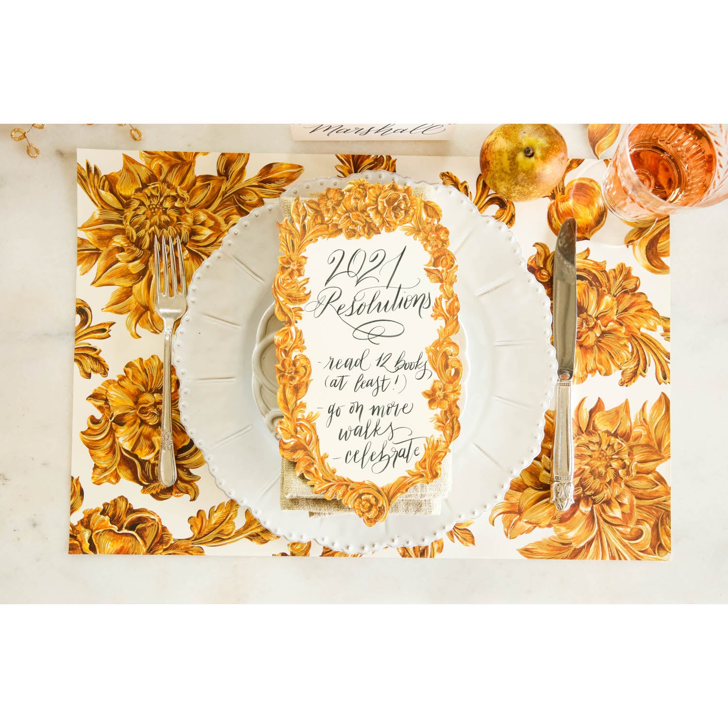 Top-down view of an elegant gold and white place setting featuring a Gold Flora Table Accent with some New Year Resolutions written on it in a beautiful script.