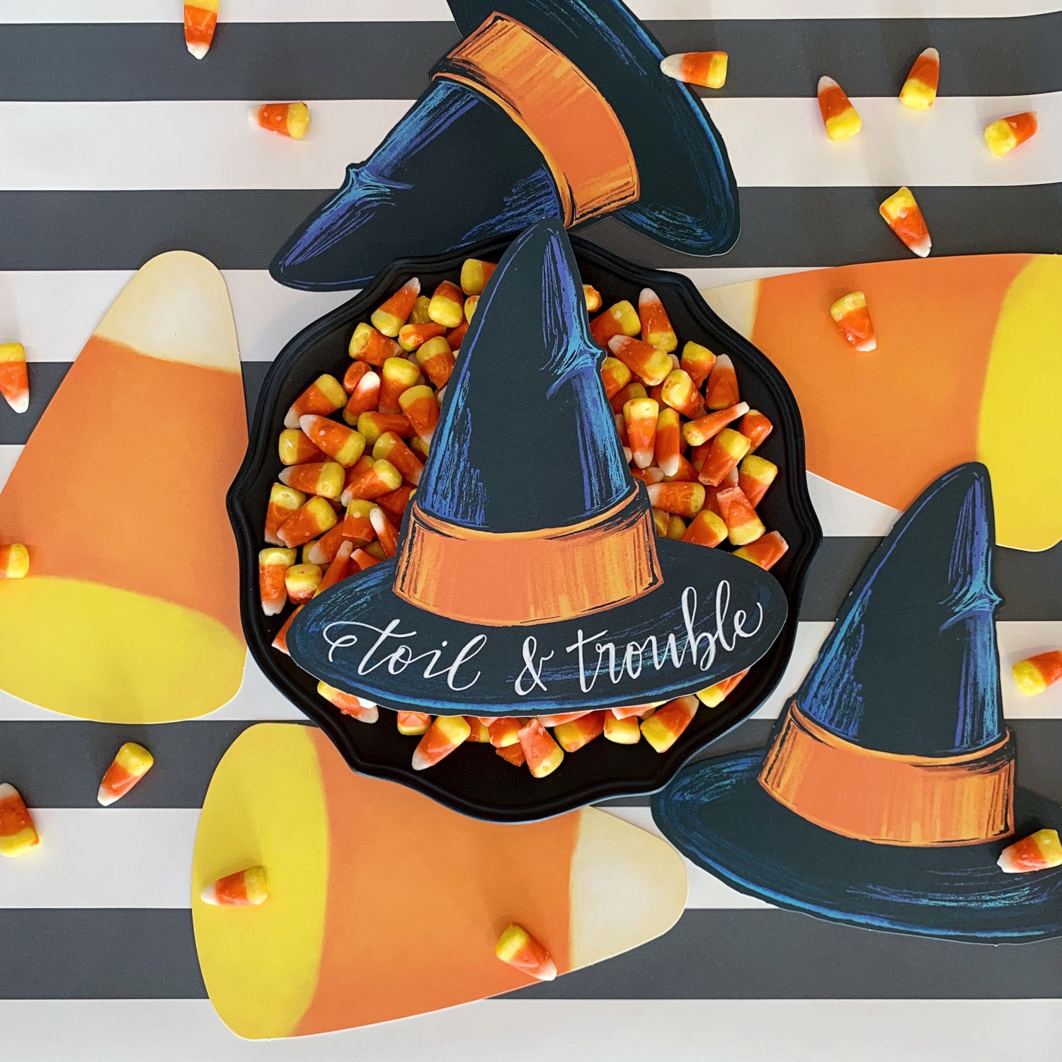 Top-down view of a Witch Hat Table Accent with "toil & trouble" written on it resting on a big bowl of candy corn, on a treat table surrounded by other Halloween table accents.