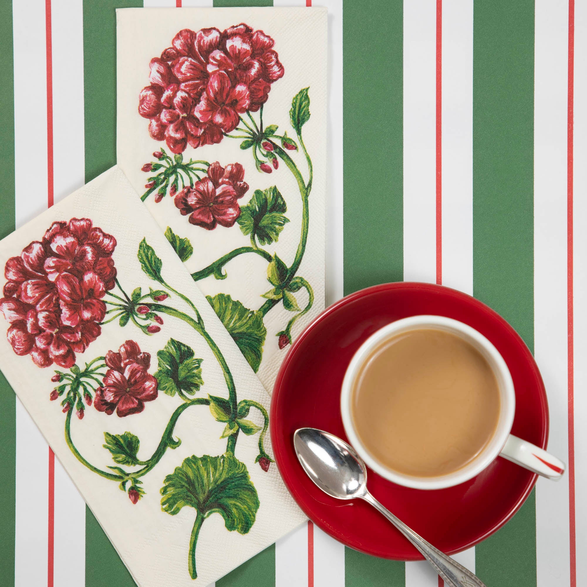 Two Geranium Guest Napkins on a red and green table runner, next to a red plate with a white up of coffee, from above.