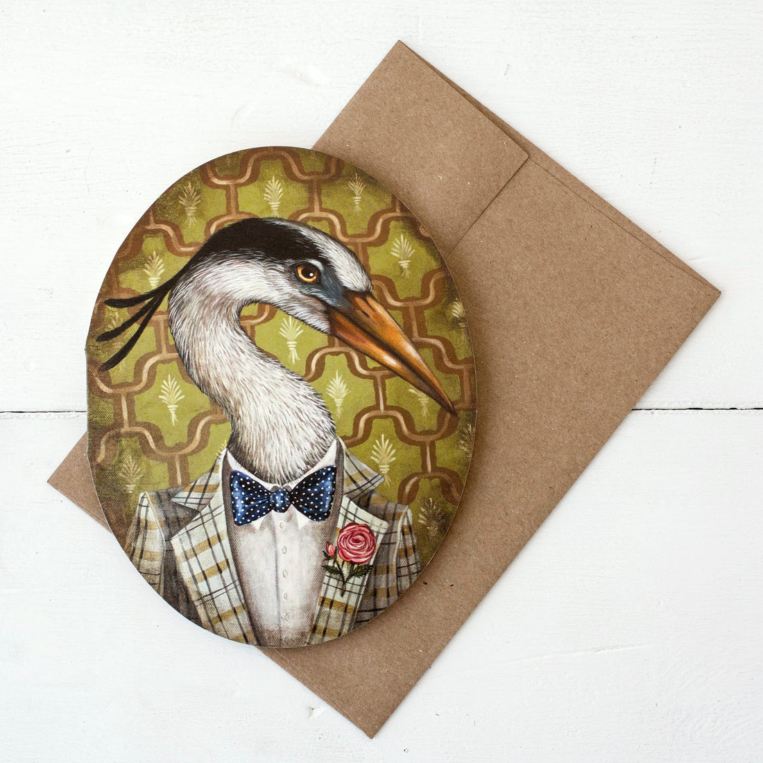 A die-cut oval greeting card with a kraft envelope, featuring a whimsical portrait of a white and black egret wearing a vintage-style suit with a red rose on his lapel over a green and orange wallpaper design.