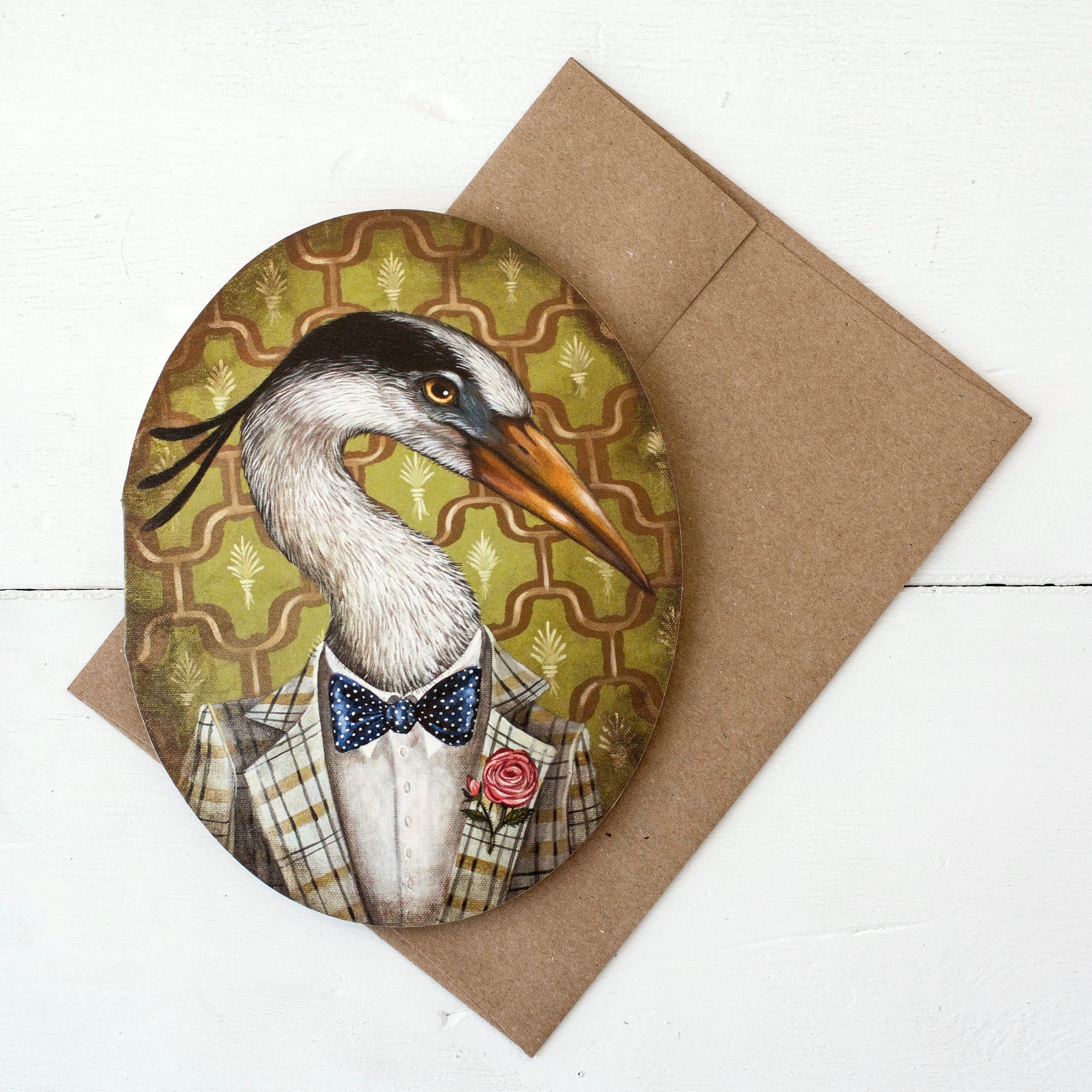 A die-cut oval greeting card with a kraft envelope, featuring a whimsical portrait of a white and black egret wearing a vintage-style suit with a red rose on his lapel over a green and orange wallpaper design.