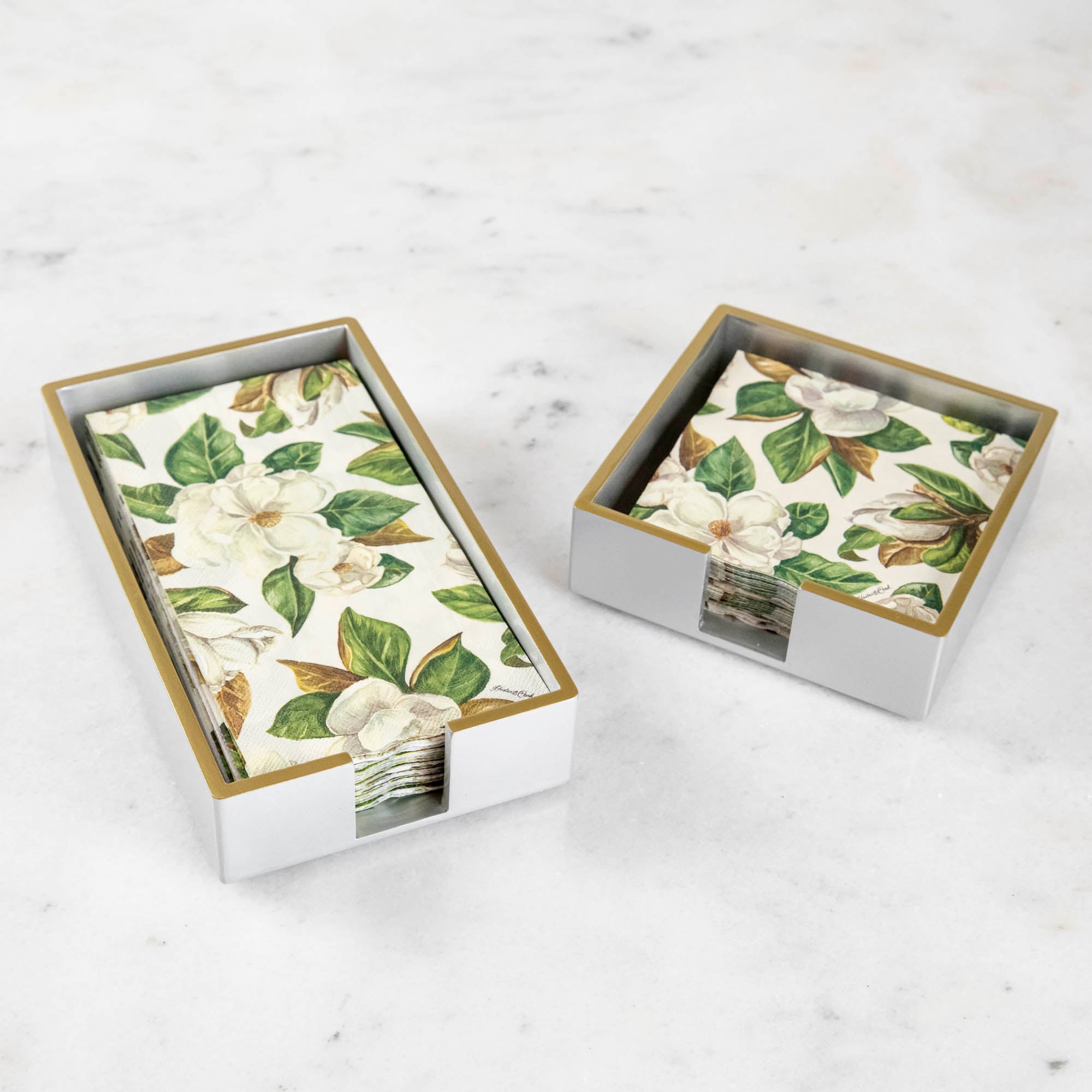 A silver colored lacquered napkin holder in guest and cocktail size with napkins in them.