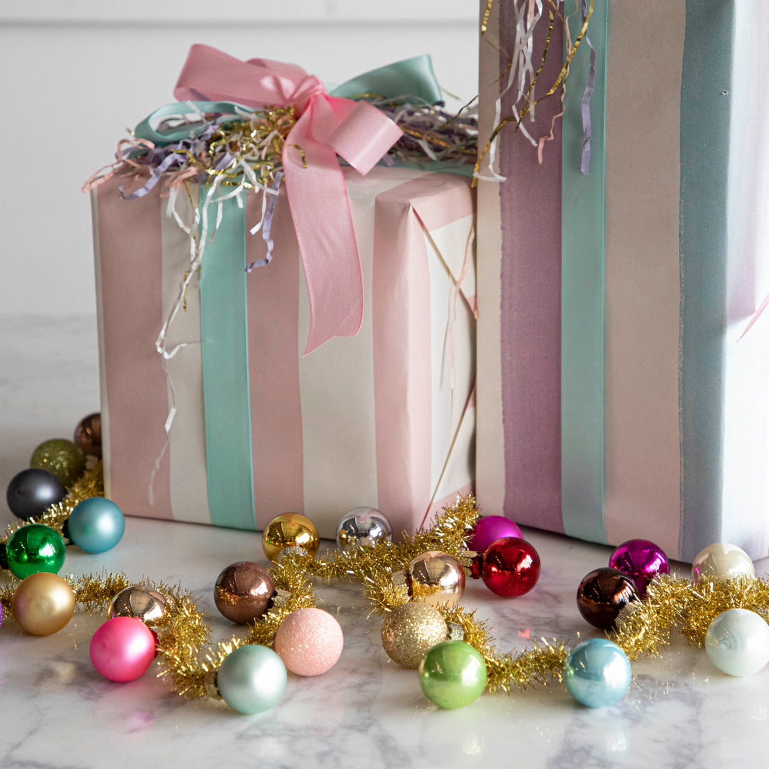 A garland made with gold tinsel string with multi colored balls attached to it strewn in front of 2 decorated gifts. 