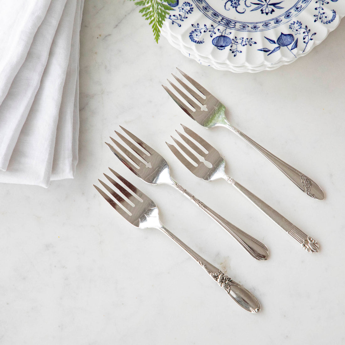 Four Hester & Cook vintage silver-plate salad forks beside a blue and white patterned plate with a white napkin and a sprig of greenery.