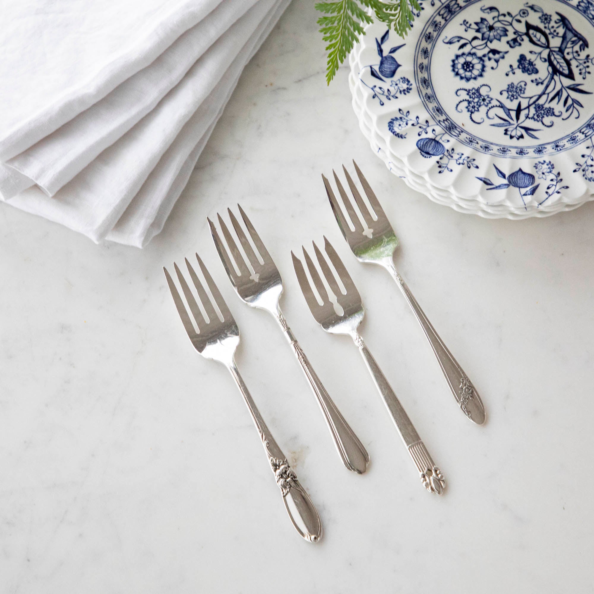 Four Hester & Cook vintage silver-plate salad forks beside a blue and white patterned plate with a white napkin and a sprig of greenery.