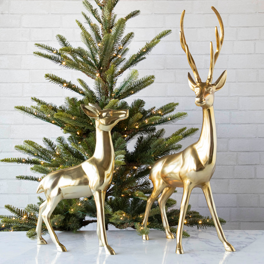 Two brass deer, a buck and a doe, on a marble table in front of a lit tree.