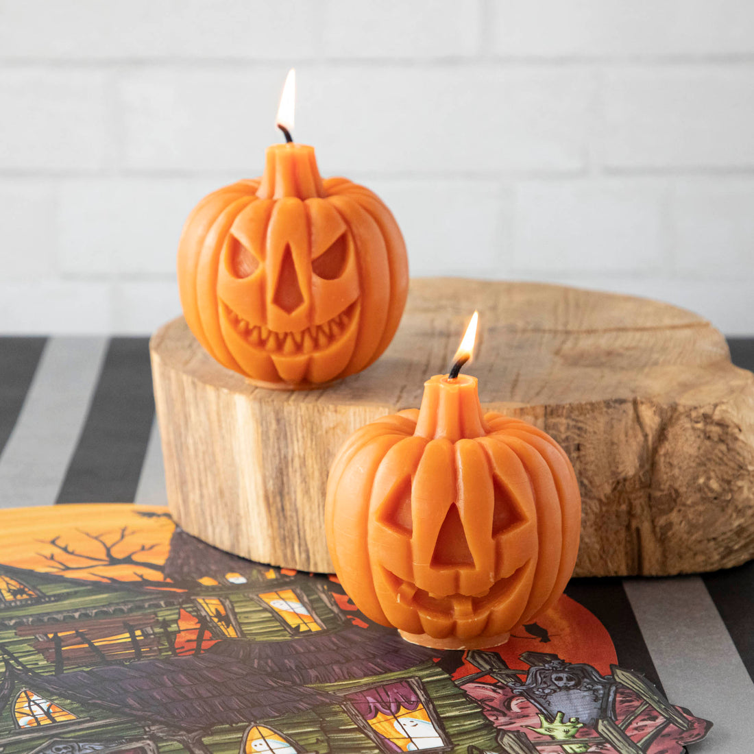 Two pumpkin-shaped candles with one side showing a scary face and the other side a happy face, lit and placed on a wooden surface.