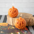 Two pumpkin-shaped candles with one side showing a scary face and the other side a happy face, lit and placed on a wooden surface.
