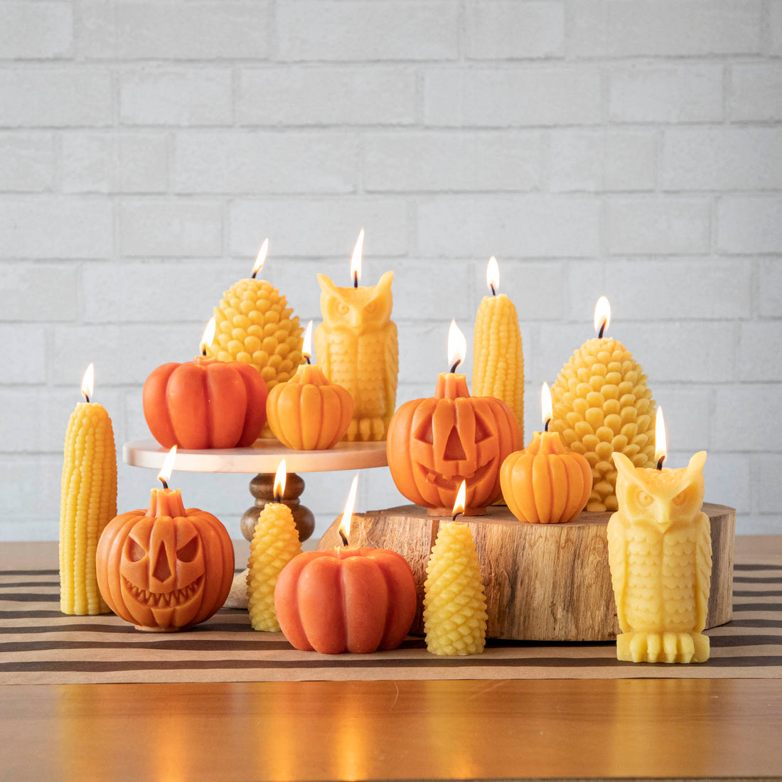A collection of pumpkin-shaped candles and other fall-themed candles, made of beeswax, displayed on a wooden surface with a few lit candles giving off a warm glow.