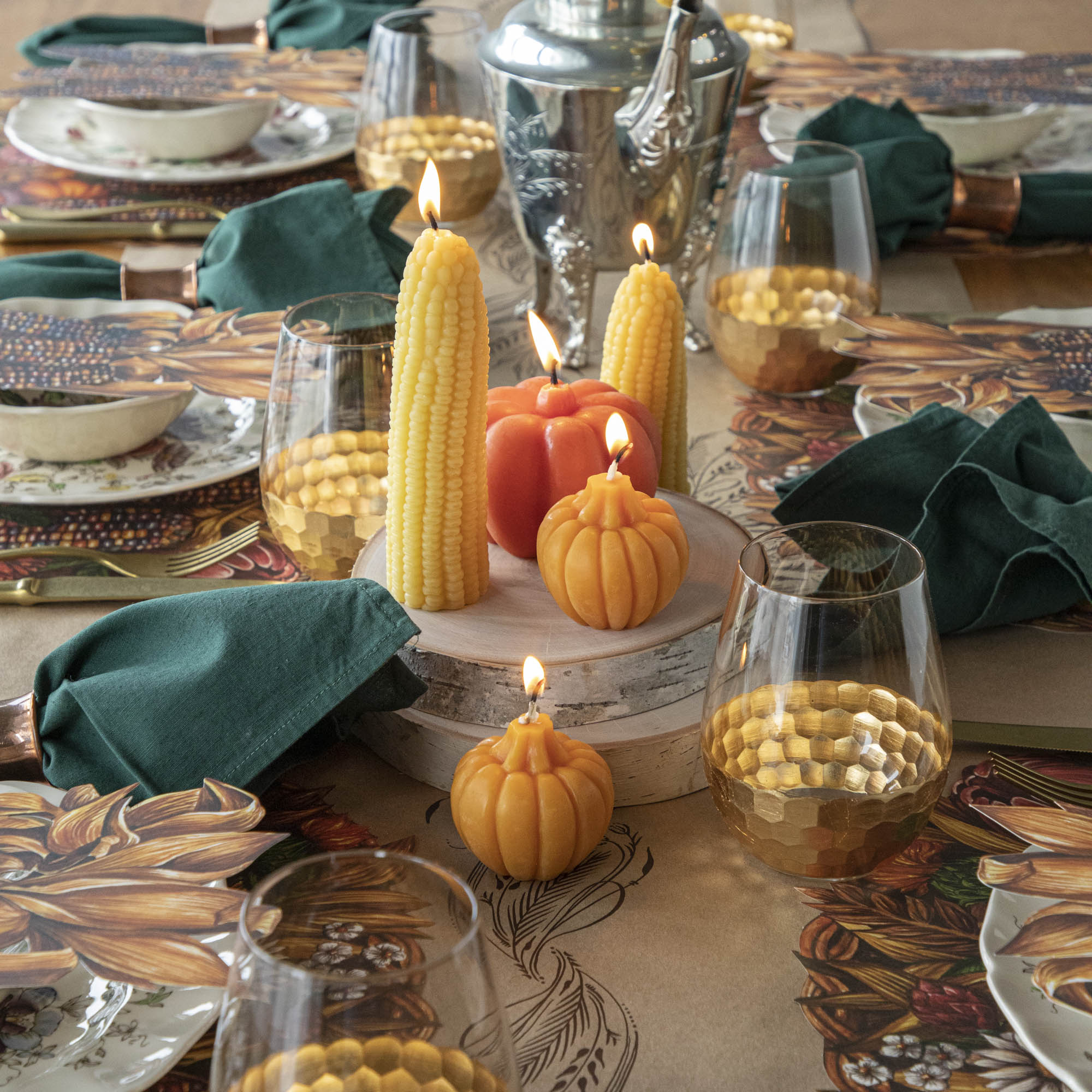 A table setting with lit beeswax corncob candles shaped like corn and pumpkins, surrounded by dishes, glasses, and green napkins.