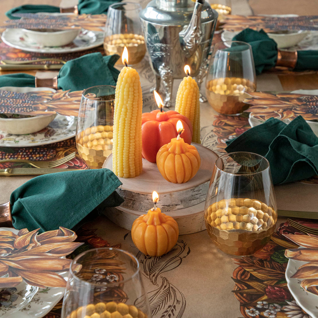 A table setting with lit beeswax corncob candles shaped like corn and pumpkins, surrounded by dishes, glasses, and green napkins.