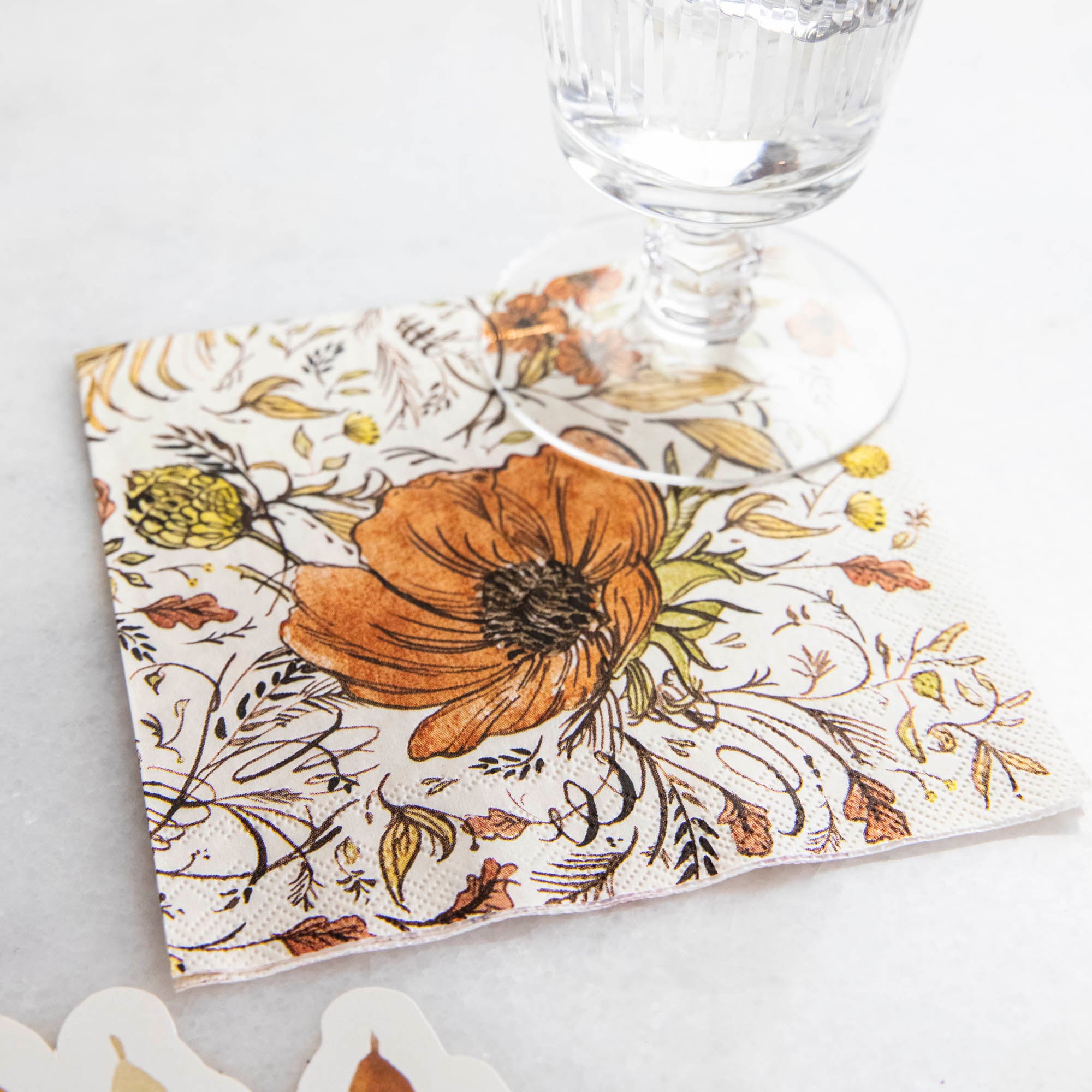 The Autumn Arrangement Cocktail Napkin on a table with a water glass sitting on it.