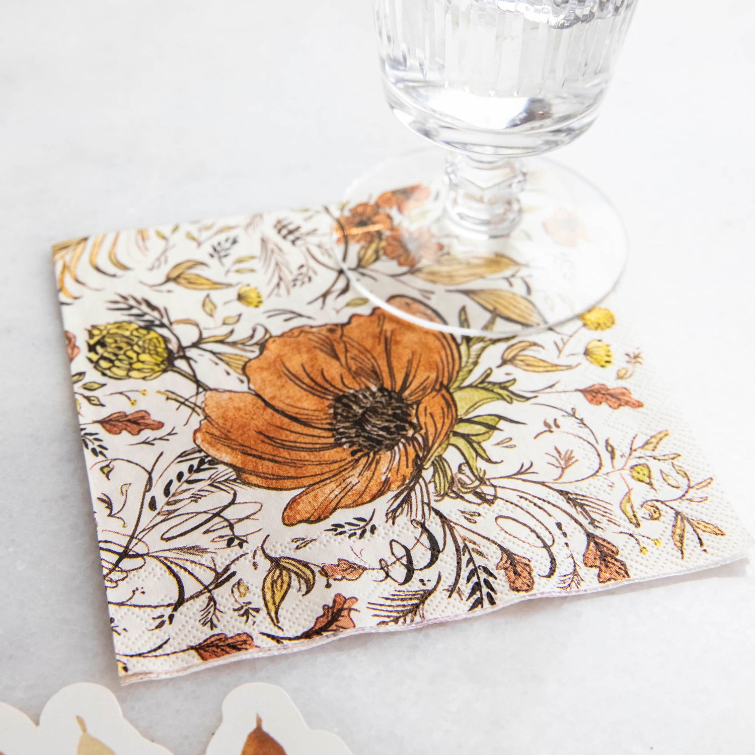 The Autumn Arrangement Cocktail Napkin on a table with a water glass sitting on it.