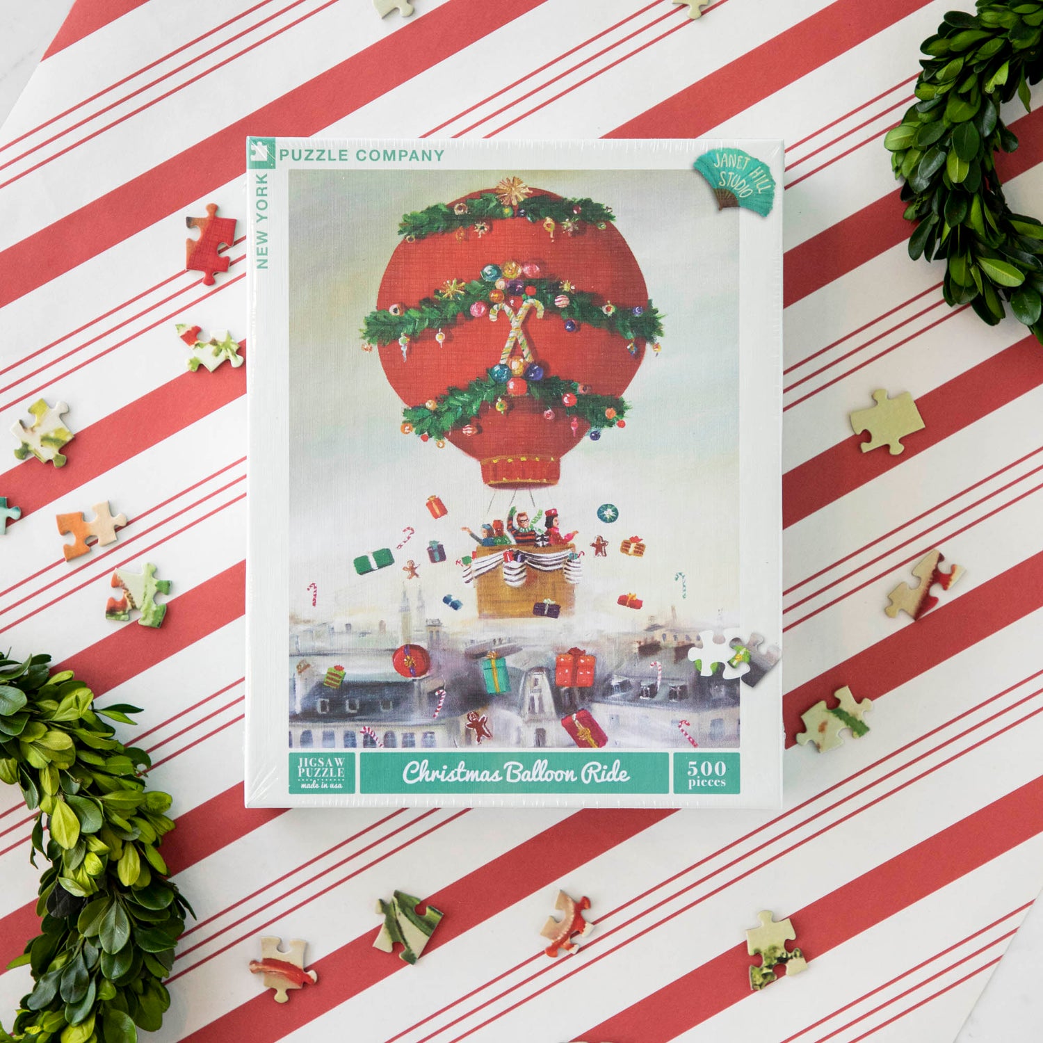 A 500 piece jigsaw puzzle depicting a whimsical painting with a Christmas theme, featuring a hot air balloon and a cityscape. The puzzle pieces are scattered around, and the puzzle has a linen style finish.