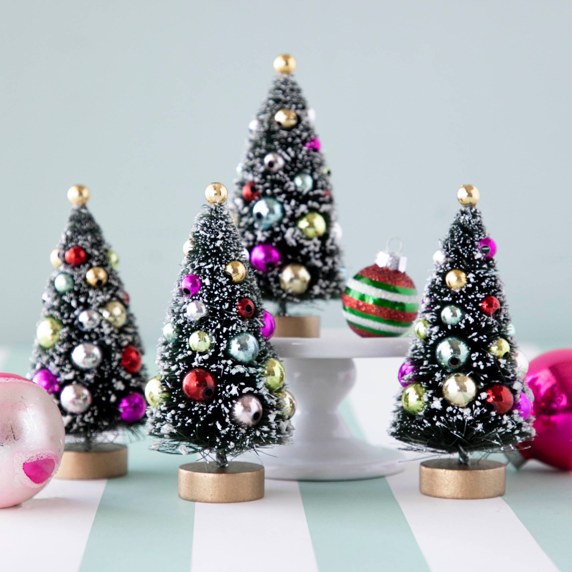 Four, 4" holiday sisal trees with colorful ornaments and snow, displayed on a table with ornaments.