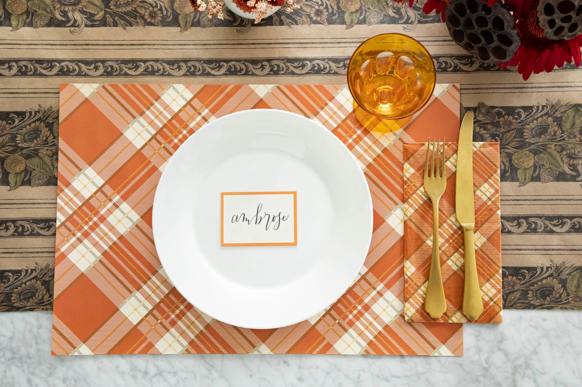 A place setting with an Autumn Arrangement placemat, Orange Plaid Runner, white plate, golden cutlery, and an amber-colored glass. The plate holds a Feather Table Accent with the name "Sophia" written on a ribbon illustration.