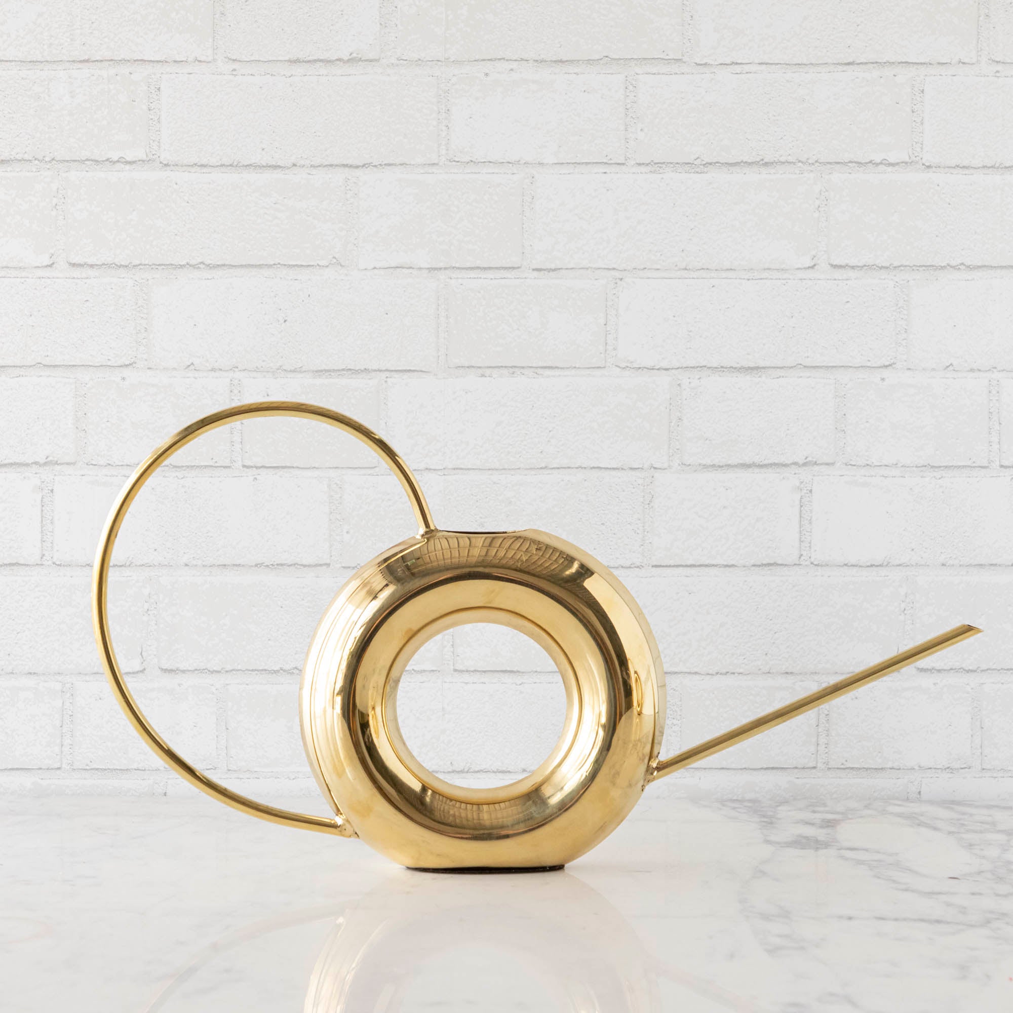 Two Accent Decor Modern Watering Cans with brass finish on a table next to flowers.