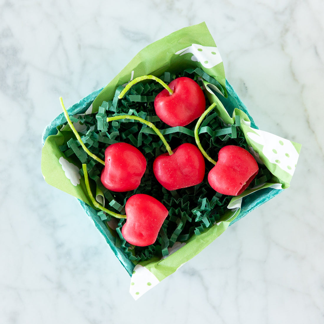 A blue carton filled with five red, cherry-shaped candles made from pure beeswax, on green paper grass, set on a white marble surface.