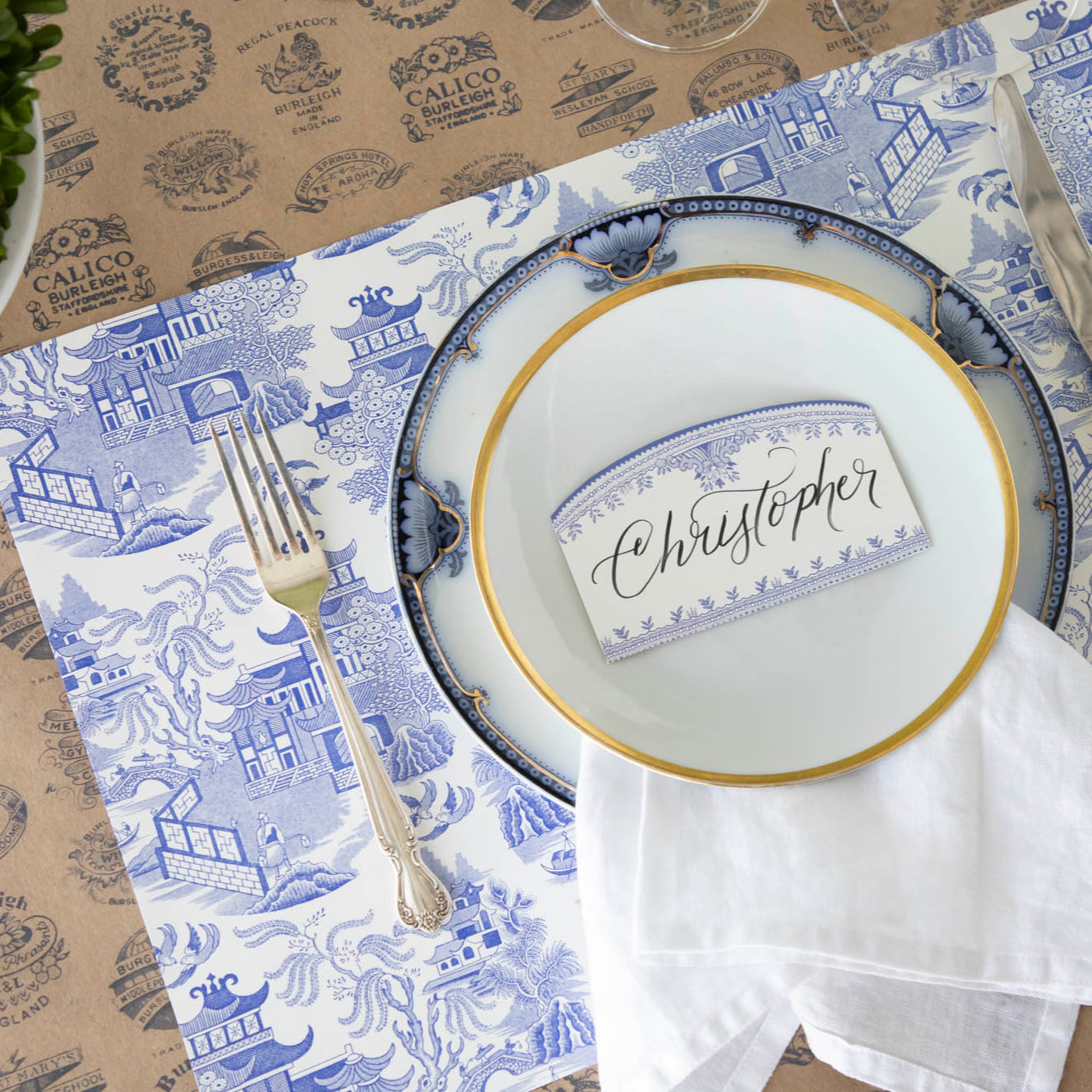 Blue Willow Place card with &quot;Christopher&quot; written in the white space, resting on dinner plates that sit on top of the Blue Willow Placemat and Kraft Burleigh Heritage Runner.