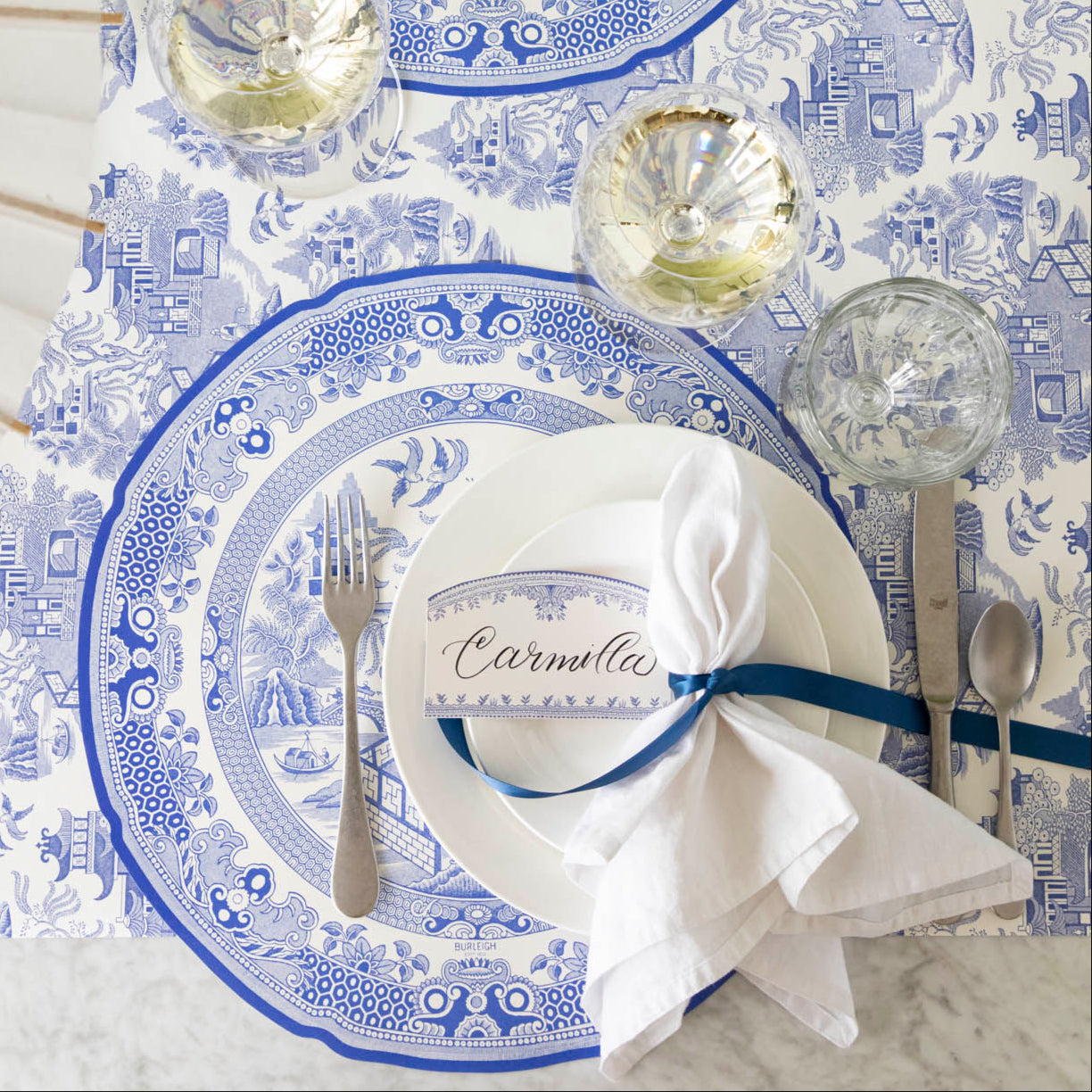Burleigh Blue Place Card with "Camilla" written in the white space, resting on dinner plates, on top of the Die-cut Blue Willow Plate Placemat. 
