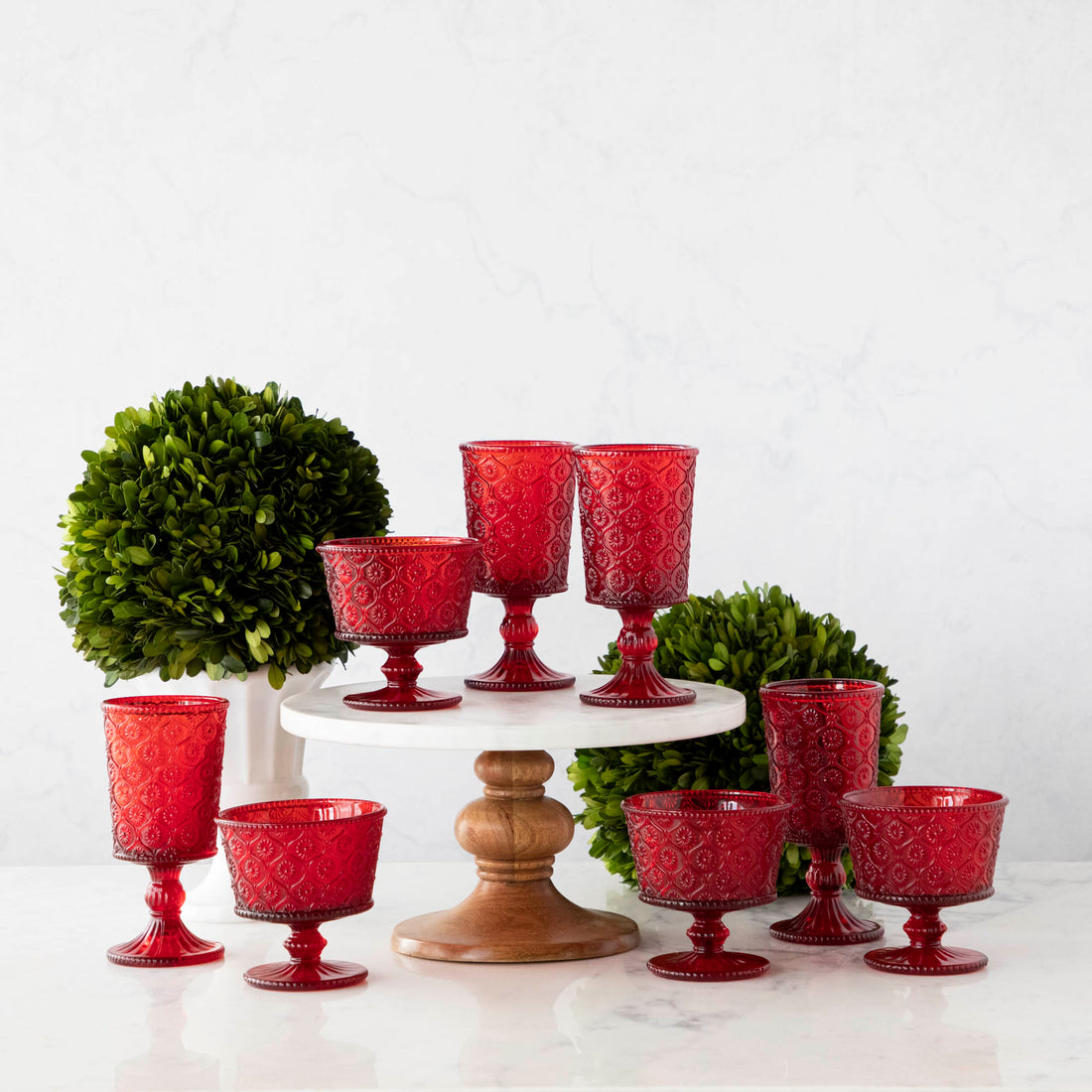 Elegant Qualia red pressed glassware set displayed on a white cake stand with wooden pedestal, flanked by green topiary balls on a marble background.