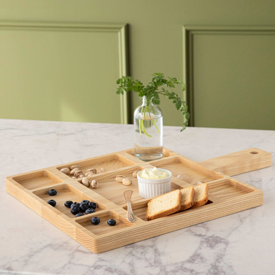 A JK Adams Ash Divided Serving Board with compartments holding pistachios, blueberries, crackers, and a dip, next to a small vase with greenery.