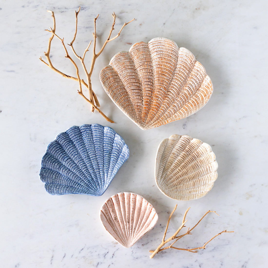 Four hand-painted stoneware shell shaped dishes in coral, peach, blue and off-white, arranged on a marble table beside branches.