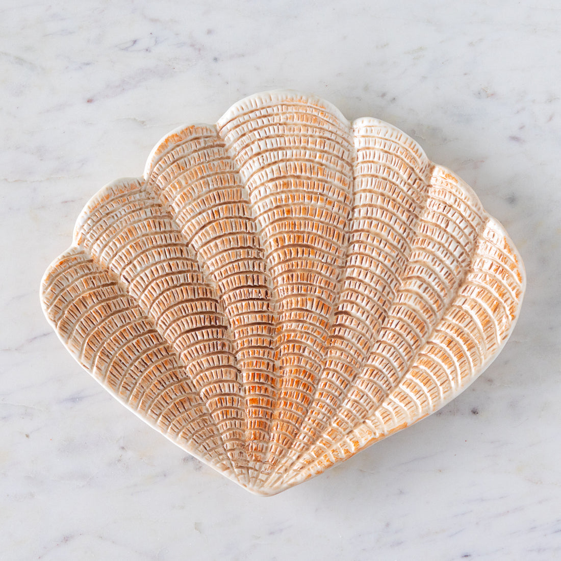 A hand-painted stoneware dish shaped like a scallop shell with textured, light peach detailing, placed on a white marble surface.