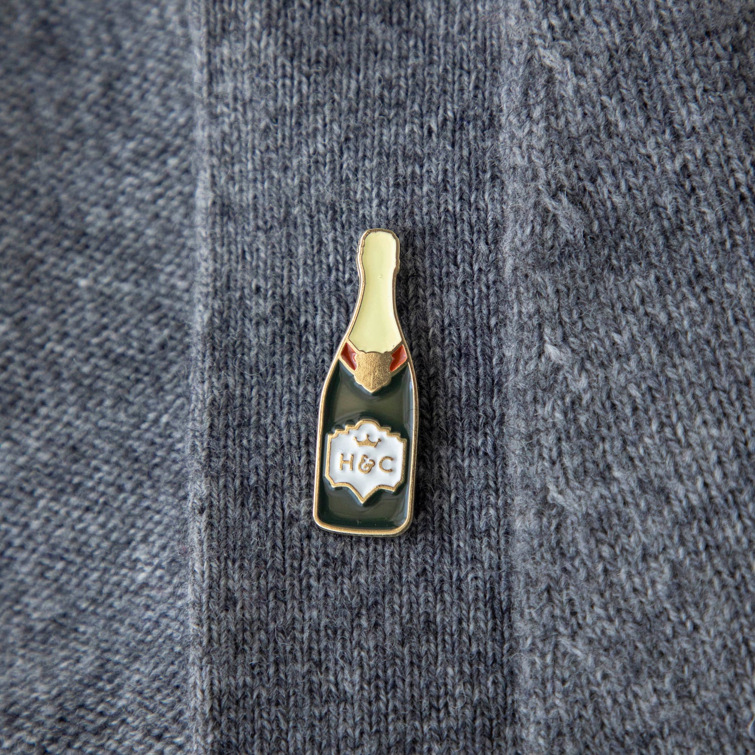 A gilded story envelops a bottle of Hester &amp; Cook Champagne Enamel Pin on a marble table.